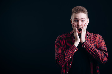 Surprised young man with wrinkled forehead touching his cheeks isolated black background copyspace