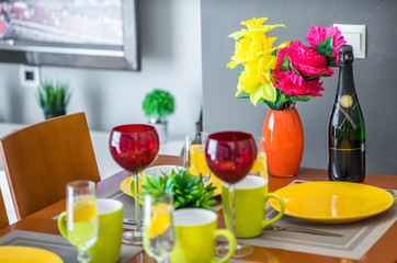 Wooden table. Yellow plates. Mugs. Water in glasses. Wine glasses. Bottle of champagne.