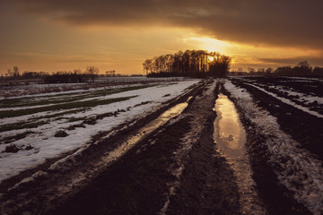 Muddy frozen rural road through fields, cloud and sunset, view in winter evening