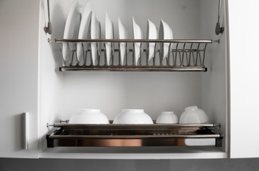 Dish drying metal rack with big nice white clean plates. Traditional comfortable kitchen. Open white dish draining closet with wet dishes of glass and ceramic, plates, bowls drying inside on rack.