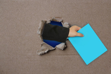 from a torn hole in an empty brown blank of cardboard, a woman’s hand holds a blue sheet of paper, the concept of an unexpected surprise