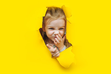A little funny red-haired girl picks her nose and squints her eyes in pleasure. Yellow background, torn paper, hole. The concept of bad behavior. Copy space.