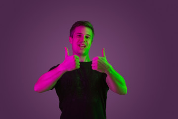 Smiling, showing thumbs up. Caucasian man's portrait on purple studio background in neon light. Beautiful male model in black shirt. Concept of human emotions, facial expression, sales, ad.