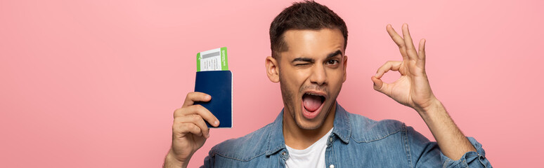 Panoramic shot of man with passport and boarding pass winking and showing okay sign isolated on pink