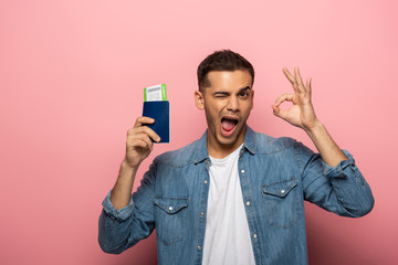 Man winking and showing okay gesture while holding passport with boarding pass on pink background