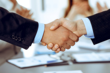 Business people shaking hands at meeting while theirs colleagues clapping and applauding. Group of unknown businessmen and women in modern white office. Success teamwork, partnership and handshake