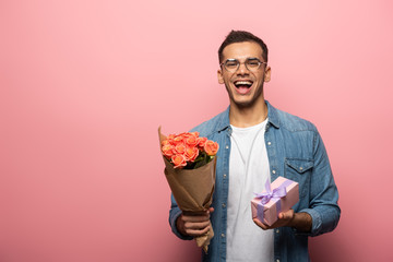 Excited man with gift and flowers looking at camera on pink background