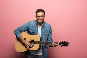 Young man smiling at camera and playing acoustic guitar on pink background