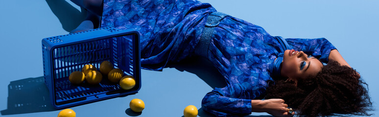 panoramic shot of african american woman lying with shopping basket and lemons on blue background