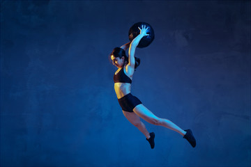 Fototapeta na wymiar Caucasian young female athlete practicing on blue studio background in neon light. Sportive model in flight, jump with ball, training. Body building, healthy lifestyle, beauty and action concept.