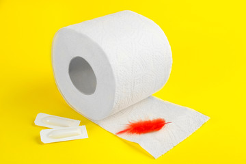 Roll of toilet paper, red feather and suppositories on yellow background. Hemorrhoid problems