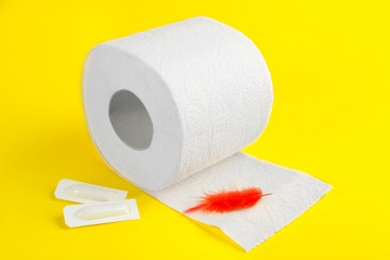 Roll of toilet paper, red feather and suppositories on yellow background. Hemorrhoid problems
