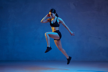 Obraz na płótnie Canvas Caucasian young female athlete practicing on blue studio background in neon light. Close up of sportive model jumping high, running. Body building, healthy lifestyle, beauty and action concept.