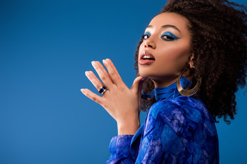 stylish african american woman in dress with makeup looking at camera isolated on blue