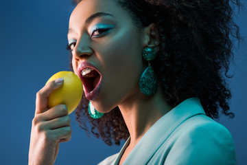 attractive african american woman with braces eating lemon isolated on blue