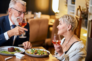 Stylish, elegant senior couple look at each other and chatting, close relationship. Grey-haired man in tuxedo raise a glass. Use e-cigarette, vape. Modern technologies