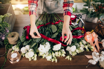 top view on cropped careful florist making, composing bouquet of white beautiful roses for customer in her own shop, red ribbon on flowers. plants, flowers, decoration concept
