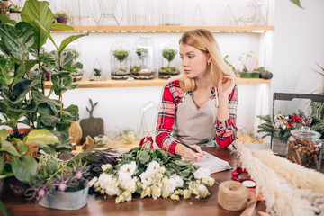 young caucasian florist lady with notebook, flowers shop owner keep records, making notes at work, wearing apron isolated in light room full off plants
