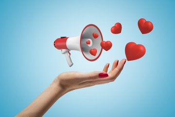Side closeup of woman's hand facing up and levitating small megaphone that emits cute red hearts, on light blue background.