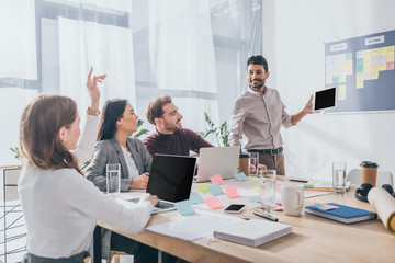 bi-racial scrum master looking at businesswoman with raising hand near coworkers