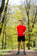 The man in a red t-shirt pose with a tennis racket and a ball on the background of green park. Sport concept