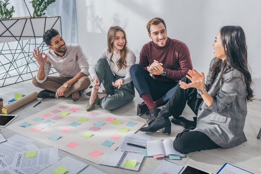 happy mixed race businessman showing ok sign near coworkers sitting on floor