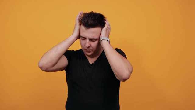 Unhappy caucasian young man wearing black shirt isolated on orange background in studio keeping hands on head, having headache. People sincere emotions, lifestyle concept.