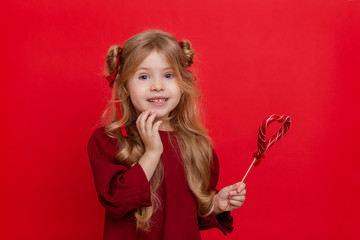 Portrait of a cheerful dreamy little girl with a heart shaped candy in her hands isolated on a red background