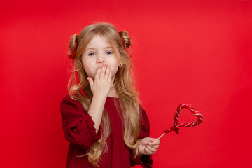 Portrait of a cheerful dreamy little girl with a heart shaped candy in her hands isolated on a red background