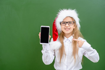 Smiling girl wearing a red christmas hat points on empty screen of the phone. Space for text