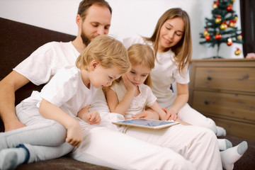 beautiful family watch video on modern tablet sitting on sofa together, dressed in white casual clothes, look at screen of tablet