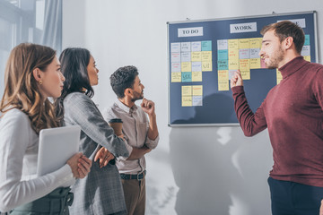 selective focus of handsome scrum master pointing with finger at board near multicultural coworkers