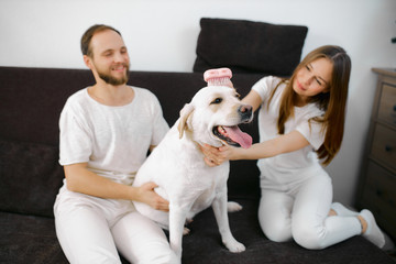lovely beautiful caucasian couple sit on sofa with their pet, white dog, wearing white casual clothes, enjoy spending time together at home, friendly dog and his owners
