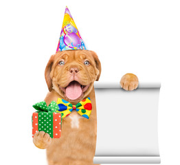 Happy puppy wearing a party hat and tie bow holds empty list and gift box. isolated on white background