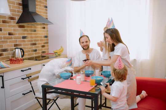 friendly caucasian family sit together in kitchen, celebrating birthday with their pet white dog, wearing birthday caps on head. people and animals concept