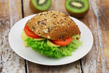 Whole grain roll with cheese, lettuce and tomato on white plate on rustic wood