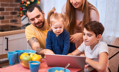 young and beautiful parents and kids watching film or video while having breakfast together in kitchen, using modern laptop, wearing casual clothes, at morning