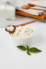 Fresh organic cottage cheese in a white bowl and a wooden spoon with sour milk cheese. Sprig of basil used as decoration. Glasses with milk or yogurt, linen napkins and cutlery in the background