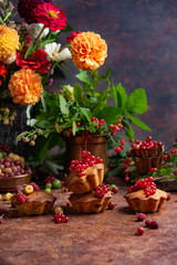 Fototapeta na wymiar Sponge cakes with berries on dark background. Chrysanthemums and greenery bunch in copper vase botanical composition. Fresh redcurrant and gooseberries on cupcakes. Home baking concept