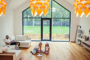 Children and parents have fun together, rest on their holiday, day off. In living room with big windows view on garden.