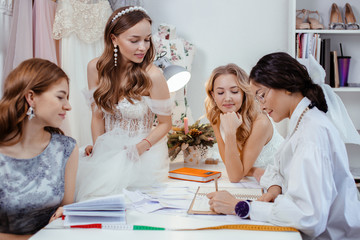 brunette adult woman designer communicate and explain which dresses are trendy, explain varieties, colors and style of dresses to young caucasian girls customers, models. isolated in wedding salon