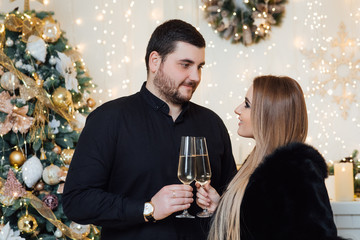 An attractive brunette girl in a black dress and a brutal man in a shirt hug on the background of a fireplace.  Man and woman banging glasses of champagne