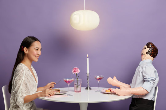 Horizontal shot of happy Korean woman enjoys time with unreal imaginary man, dreams about perfect relationships, has fake love, waits for boyfriend at festive dinner table with male doll. Loneliness