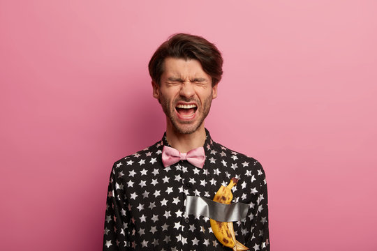 Emotive hipster with trendy hairstyle, dressed in fashionable black shirt with bowtie, wears duct taped banana peel as modern trend, poses against pink background. Art installation, popular picture