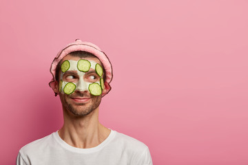 Good tempered happy man wears face mask and cucumbers, enjoys morning spa procedure, wants to look...