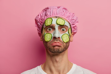 Horizontal shot of unhappy man gets cosmetology treatments, refreshes skin with vegetable mask, stares at camera, wears bath hat, poses against pink background. Cosmetology, skin care concept