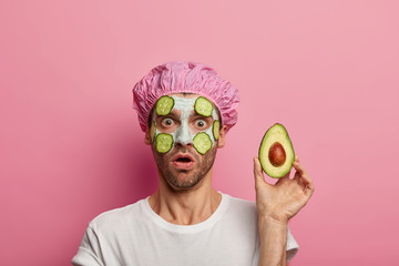 Photo of stunned man gasps from wonder, holds avocado near face, surprised by effect of clay mask, wears bath cap, has bristle, isolated over pink studio wall. Beauty, wellness, skin care, cosmetology
