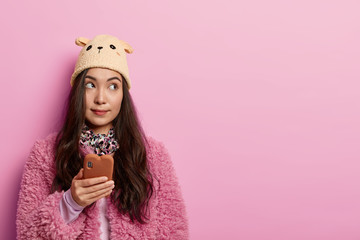 Horizontal view of pensive woman with dark hair uses mobile phone for chatting online, thinks about which comment to write under post, stands with modern gadget indoor, copy space on rosy wall