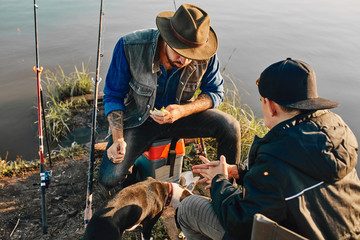 Caucasian adult father and teen son sit on fishing chairs and eat sandwich after good fishing. They tired, but happy