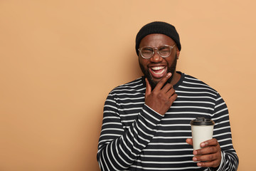 People, lifestyle, emotions concept. Joyful black man holds chin, smiles sincerely, drinks takeout...